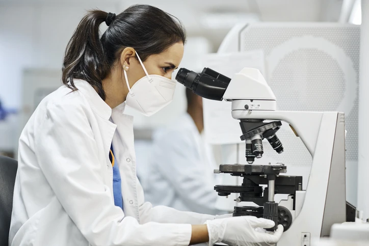 Cientista feminina de jaleco e máscara realizando análise laboratorial com microscópio profissional em ambiente de pesquisa científica Cientista usando microscópio em laboratório de pesquisa