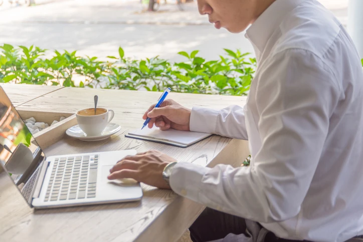 Homem de camisa social trabalhando remotamente em uma cafeteria com notebook e caderno para produtividade profissional