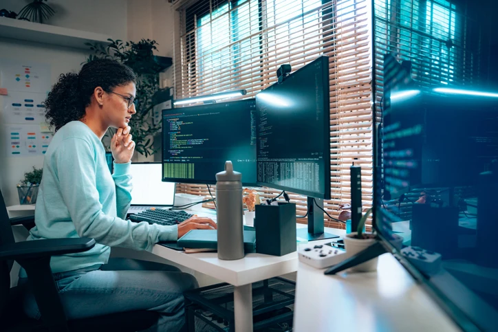 Programadora trabalhando em casa com múltiplos monitores focada em codificação