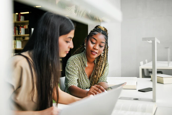 Estudantes universitárias colaborando em um projeto acadêmico usando notebook em uma biblioteca silenciosa