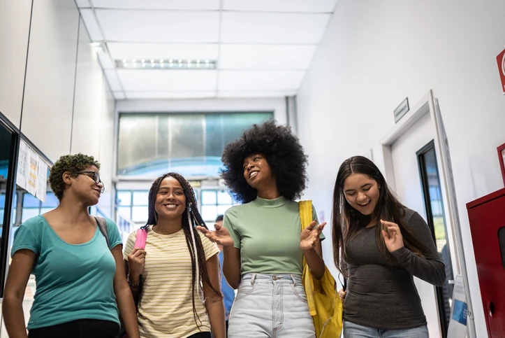 Estudantes universitárias diversas caminhando e conversando animadamente em um corredor de uma instituição de ensino superior