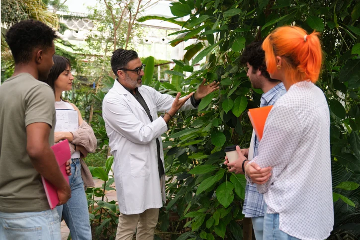 Professor em jaleco branco explica características de plantas para grupo de estudantes universitários em aula de campo em estufa