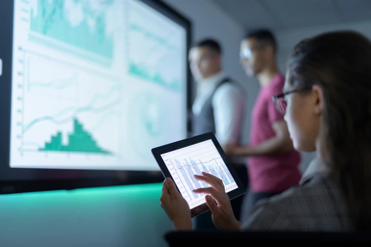 Equipe profissional analisando gráficos de dados financeiros em tablet e tela grande
