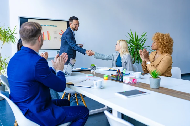 Equipe corporativa aplaudindo um aperto de mão entre um gestor e uma colaboradora durante uma reunião de resultados.