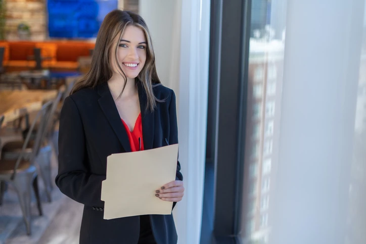 Profissional de negócios sorridente pronta para uma reunião segurando documentos importantes em um escritório corporativo Mulher jovem profissional sorridente segurando uma pasta de documentos em um ambiente de escritório moderno