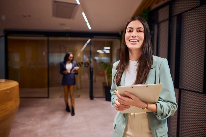 Profissional sorridente segurando uma prancheta em um ambiente corporativo moderno e colaborativo Mulher sorridente segurando uma prancheta em um ambiente de escritório moderno