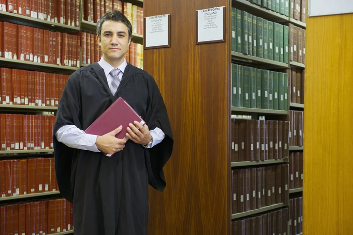 Advogado ou estudante de direito vestindo toga acadêmica segurando um livro em uma biblioteca jurídica