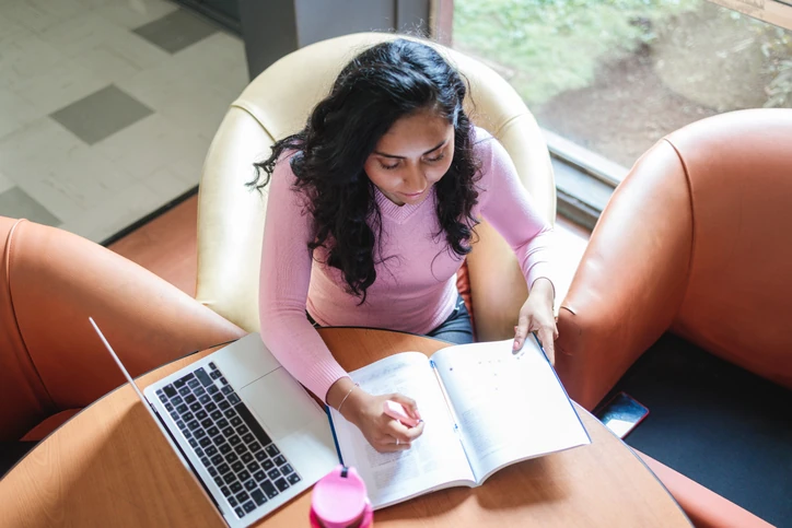 Estudante universitária concentrada nos estudos utilizando notebook e material didático em um ambiente acadêmico Estudante focada escrevendo em caderno com notebook ao lado em ambiente de estudo