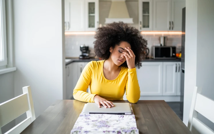 Mulher jovem e negra com cabelo cacheado sentada à mesa da cozinha, demonstrando exaustão e estresse enquanto trabalha de casa com um laptop fechado.