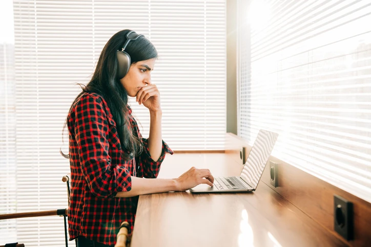 Estudante concentrada utilizando notebook e fones de ouvido para estudos online ou trabalho remoto em um ambiente iluminado Estudante focada usando notebook e fones de ouvido em ambiente de estudo