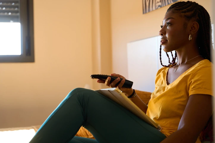 Jovem mulher negra com tranças relaxando no quarto segurando um controle remoto