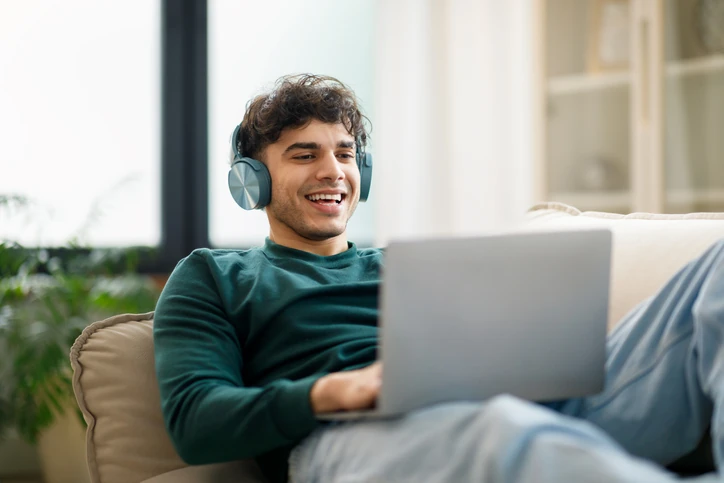 Jovem estudante sorrindo enquanto usa notebook e fones de ouvido em um sofá confortável