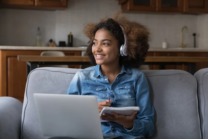 Jovem estudante sorridente sentada no sofá usando fones de ouvido e caderno para estudos online