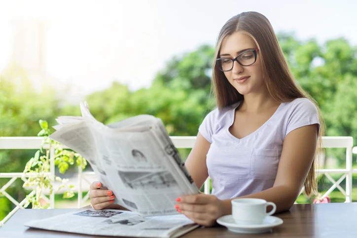 Jovem estudante focada lendo jornal matinal e tomando café em um ambiente externo tranquilo para um momento de lazer Jovem estudante lendo jornal em uma mesa ao ar livre com xícara de café