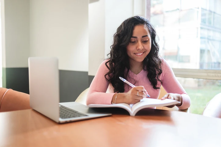 Estudante sorrindo escrevendo em um caderno ao lado de um notebook em uma mesa