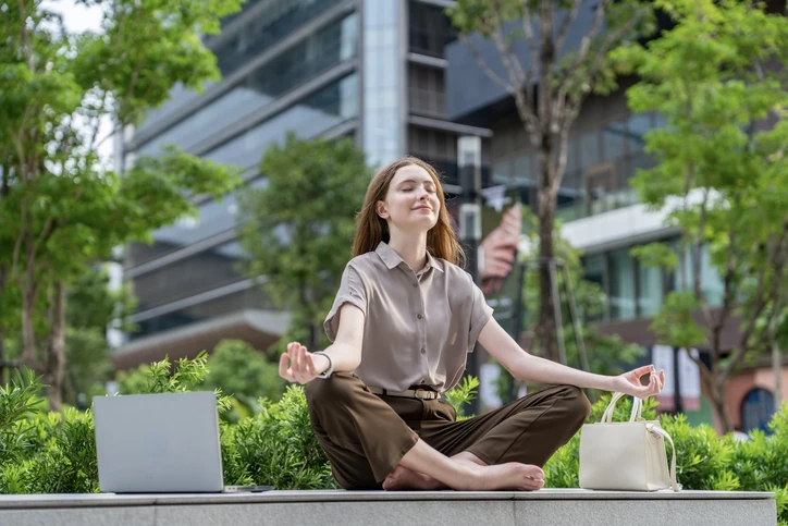 Equilíbrio entre vida profissional e pessoal: mulher praticando meditação ao lado de notebook em área urbana corporativa Mulher jovem praticando meditação e mindfulness em um ambiente corporativo ao ar livre