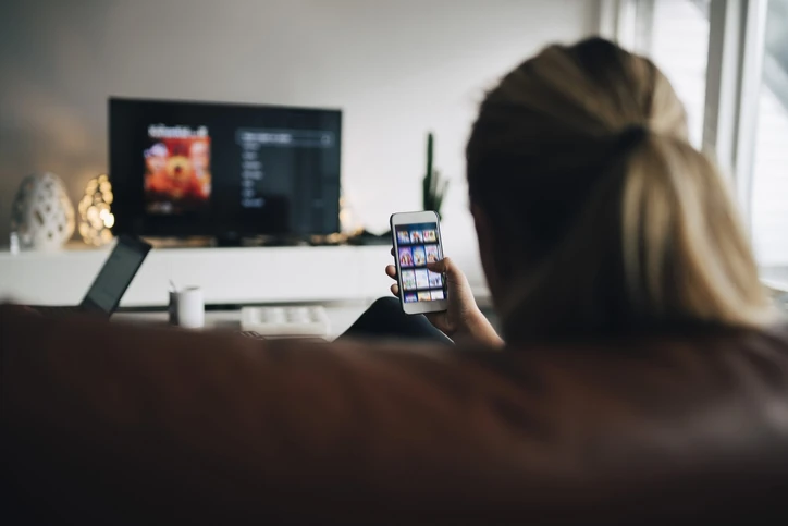 Mulher utilizando aplicativo de streaming no celular enquanto assiste televisão confortavelmente em sua casa Mulher relaxando no sofá usando smartphone enquanto assiste TV na sala de estar