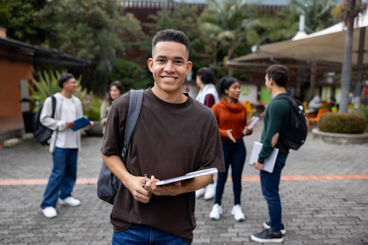 Estudante universitário sorridente segurando material de estudo no campus da faculdade com colegas de turma interagindo ao fundo