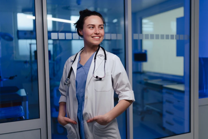Médica profissional sorridente em ambiente de hospital moderno pronta para atendimento clínico e cuidados de saúde Jovem médica sorridente usando jaleco branco e estetoscópio em um ambiente hospitalar