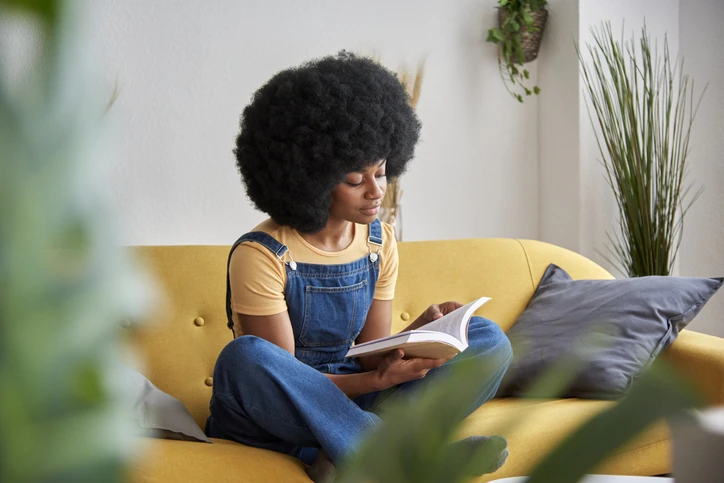 Jovem mulher negra com cabelo afro sentada em um sofá amarelo lendo um livro em um ambiente aconchegante