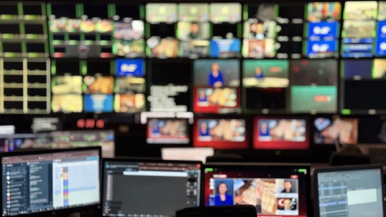 Bastidores da produção de televisão em uma sala de controle técnica com múltiplos monitores de edição e transmissão ao vivo
