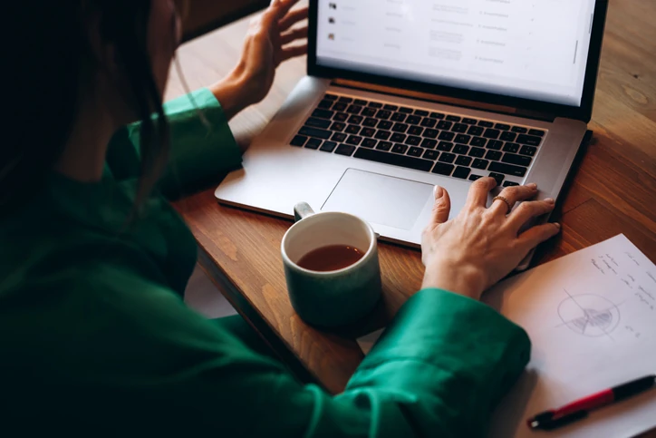 Pessoa trabalhando em um laptop com uma xícara de café e documentos sobre uma mesa de madeira