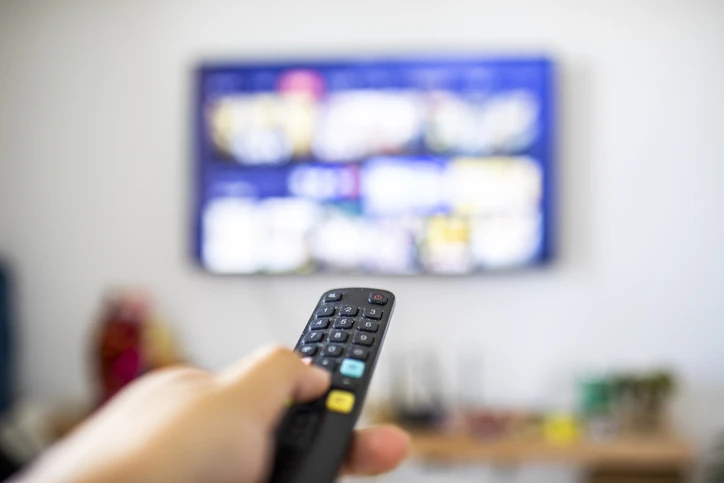 Mão segurando controle remoto apontado para uma televisão com tela desfocada