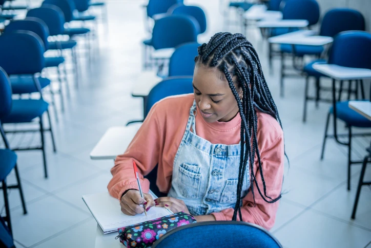 Estudante negra com tranças escrevendo em um caderno em uma sala de aula vazia