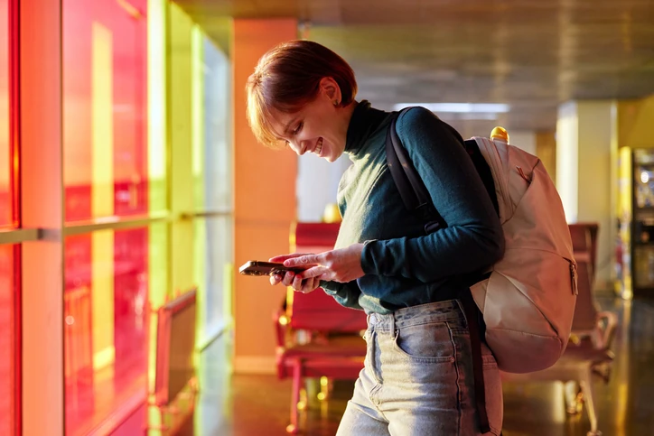 Estudante universitária utilizando smartphone em ambiente de campus ou aeroporto com iluminação vibrante