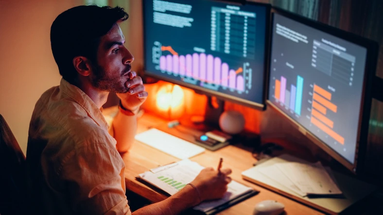 Homem analisando gráficos de dados financeiros em monitores durante a noite