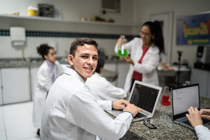 Estudante de ciência sorrindo enquanto usa notebook em laboratório de química com professora e colegas ao fundo