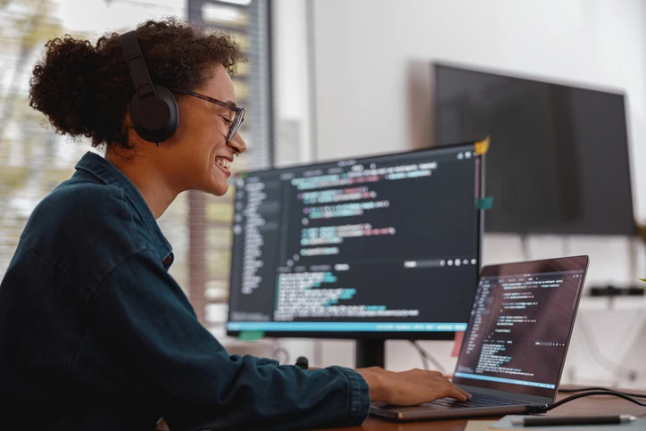 Programadora negra sorrindo enquanto trabalha em código no computador com fones de ouvido