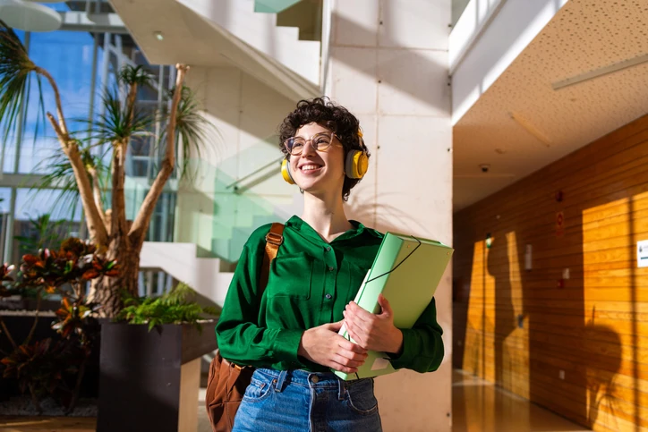 Estudante universitária sorridente usando fones de ouvido e segurando pastas em um campus moderno