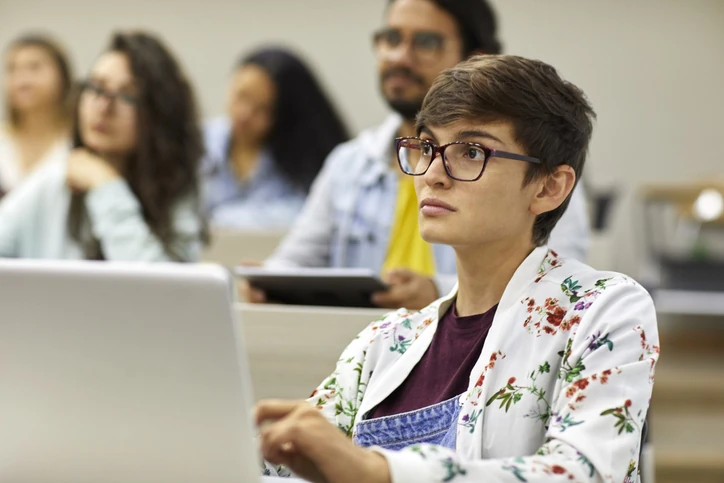 Estudante atenta usando notebook em sala de aula universitária com outros alunos ao fundo