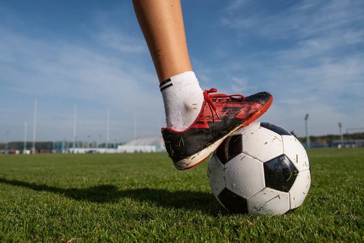 https://estaticos.animaeducacao.com.br/medias/20260622102945/img-blog-alem_dos_gramados.webp Pé de um jogador de futebol com chuteira apoiado sobre uma bola de futebol em um campo gramado
