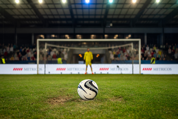 Visão de perto de uma bola de futebol posicionada no centro do campo antes de um pênalti com goleiro desfocado ao fundo