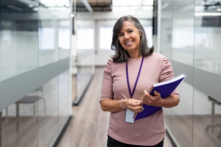 Mulher sorrindo, segurando livros e cadernos, em um ambiente corporativo, representando uma profissional da área da educação e gestão.