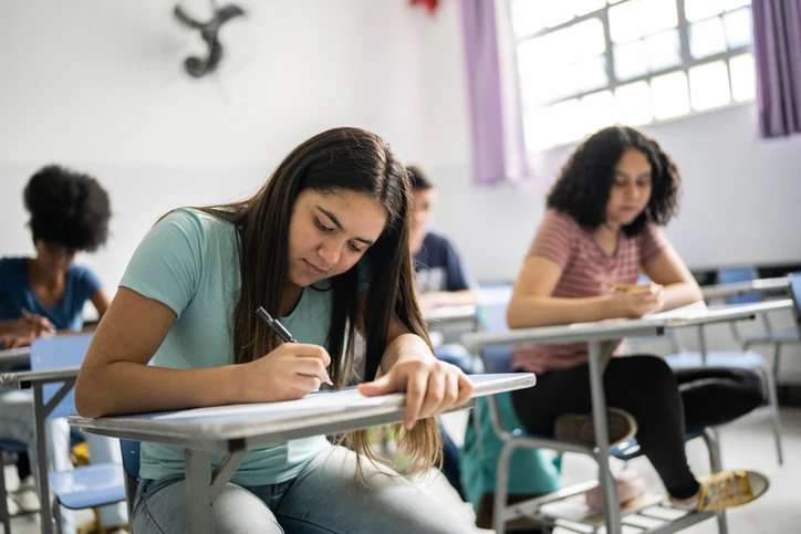 Estudante escreve em uma folha durante uma atividade em sala de aula, enquanto outros alunos realizam suas tarefas ao fundo. Estudante escreve em uma folha durante uma atividade em sala de aula, enquanto outros alunos realizam suas tarefas ao fundo.