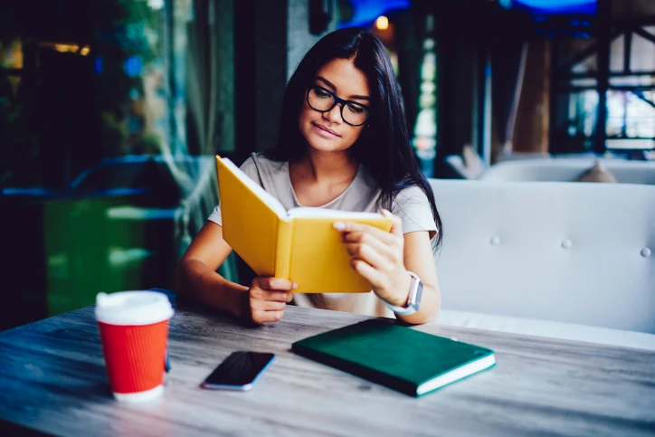 : Jovem mulher de óculos lendo um livro amarelo em uma cafeteria, sentada à mesa com um celular, uma xícara de café e outro livro.