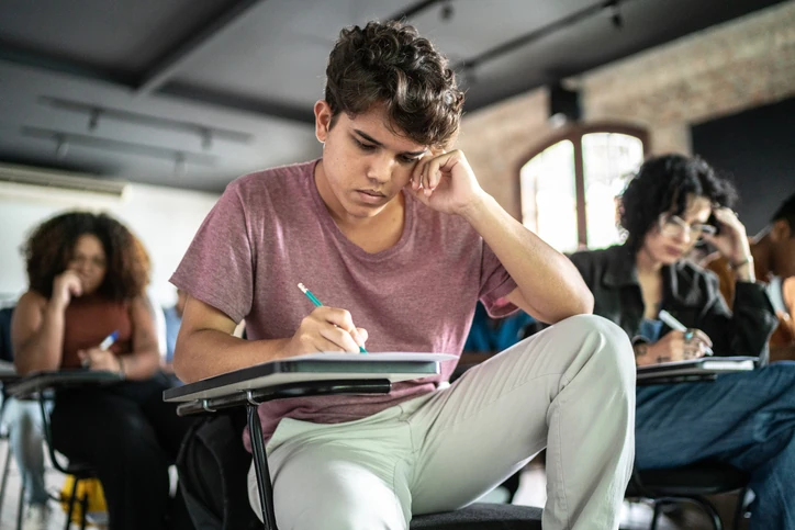Estudante sentado em uma sala de aula, escrevendo em um caderno sobre a carteira enquanto apoia a cabeça em uma das mãos; outros estudantes aparecem ao fundo. Estudante sentado em uma sala de aula, escrevendo em um caderno sobre a carteira enquanto apoia a cabeça em uma das mãos; outros estudantes aparecem ao fundo.