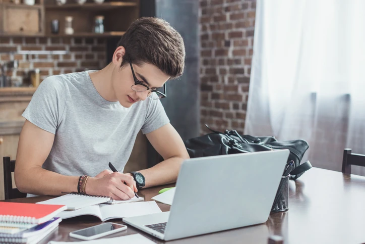 estudante-preparacao-enem-primeira-vez Jovem estudante fazendo anotações em um caderno, com um notebook aberto à frente, se preparando para o ENEM.