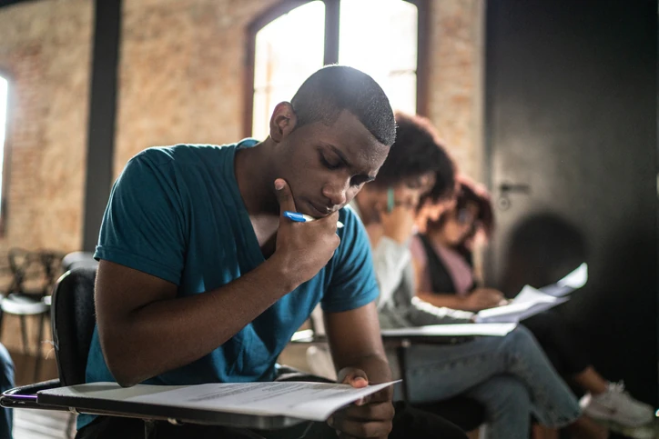 Estudante sentado em uma carteira, escrevendo em uma folha enquanto segura uma caneta junto ao queixo; outros estudantes aparecem desfocados ao fundo.