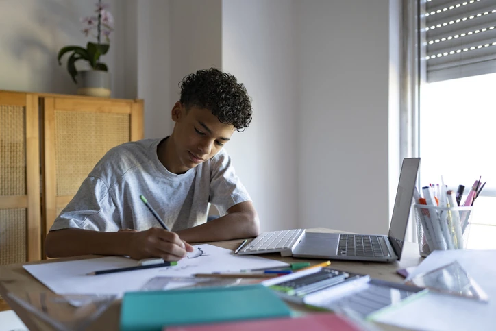 Estudante adolescente estudando para uma prova em casa, escrevendo em uma folha ao lado de livros e notebook.