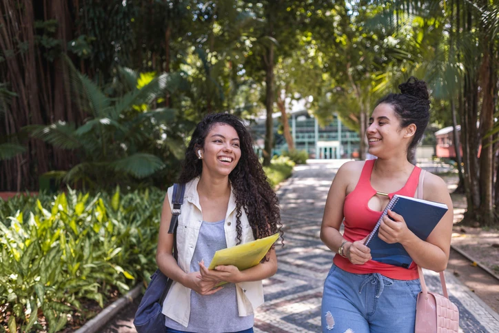 Duas jovens caminham sorrindo e conversando em um campus cercado por muita vegetação.