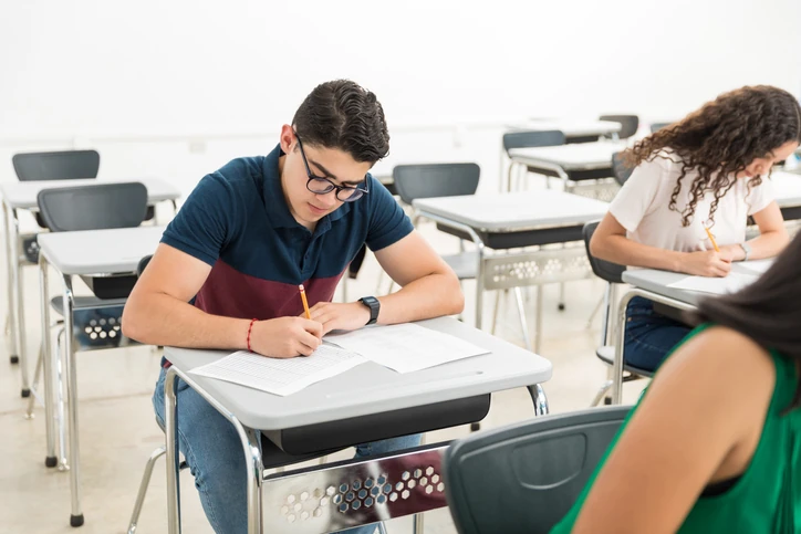 Estudante realizando uma prova escrita em sala de aula, com outros alunos ao fundo.
