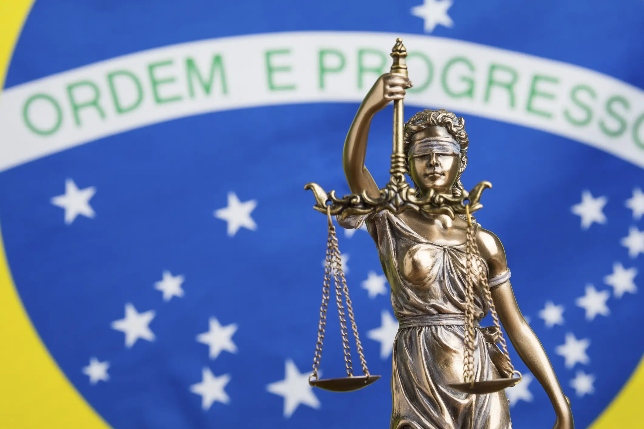 Estátua da deusa Têmis, símbolo da justiça, com a bandeira do Brasil ao fundo.