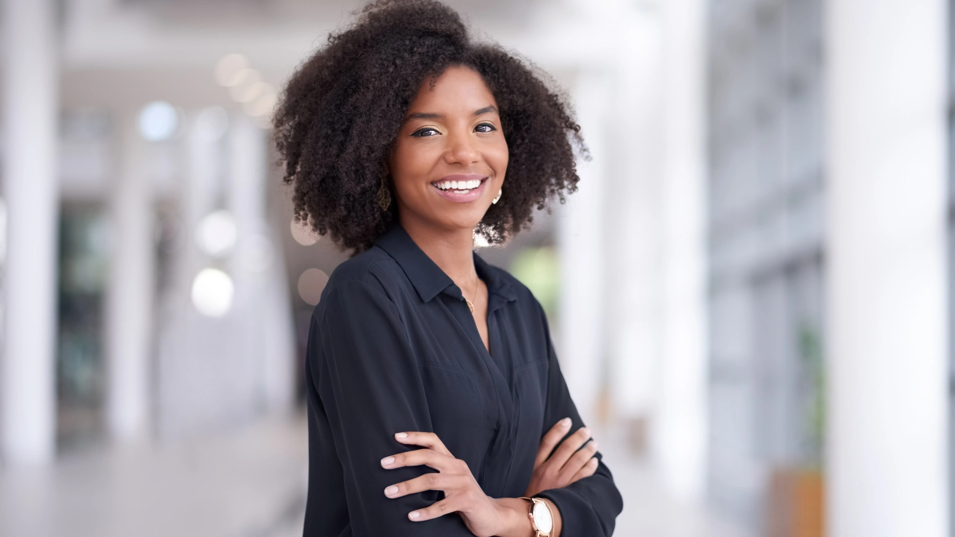 Jovem mulher negra sorridente com cabelo cacheado volumoso usando camisa preta em ambiente corporativo