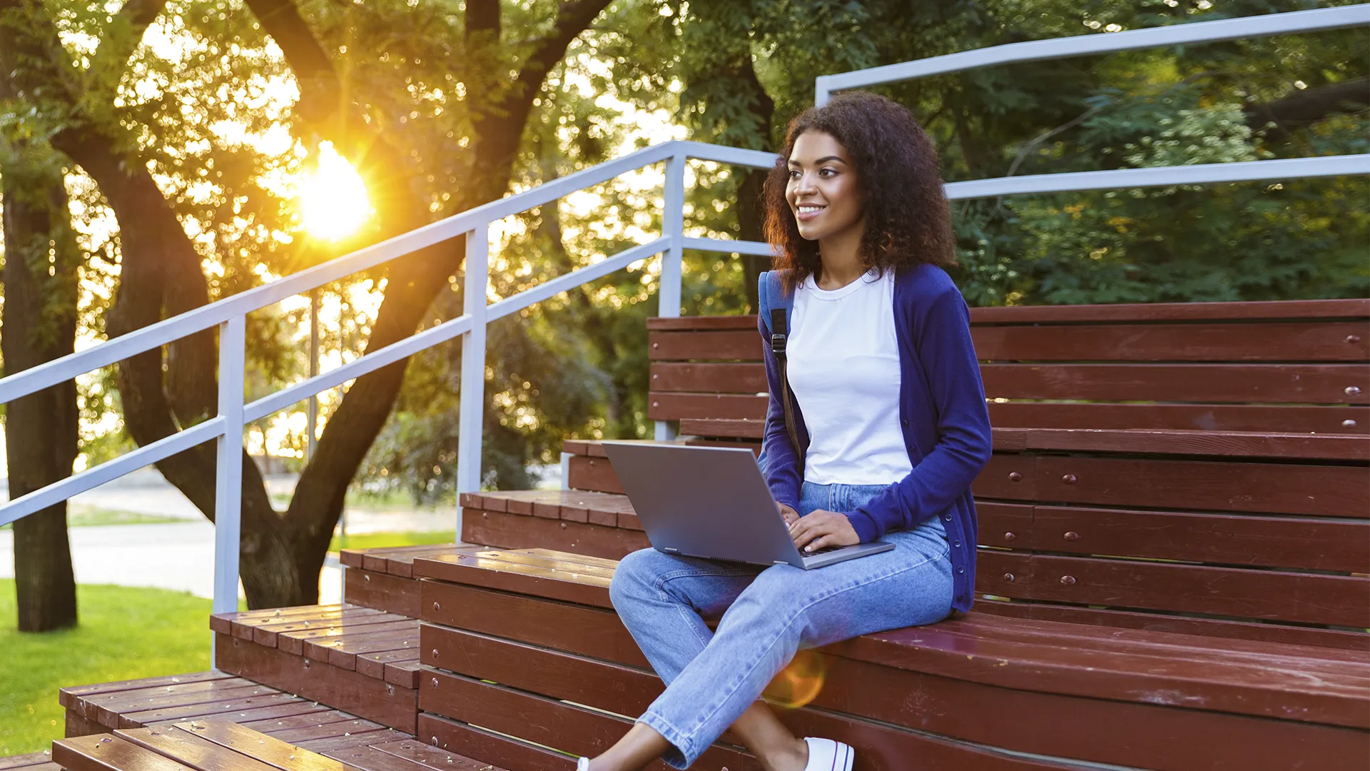 Estudante jovem e sorridente usando notebook sentada em escadaria de madeira ao ar livre durante o pôr do sol