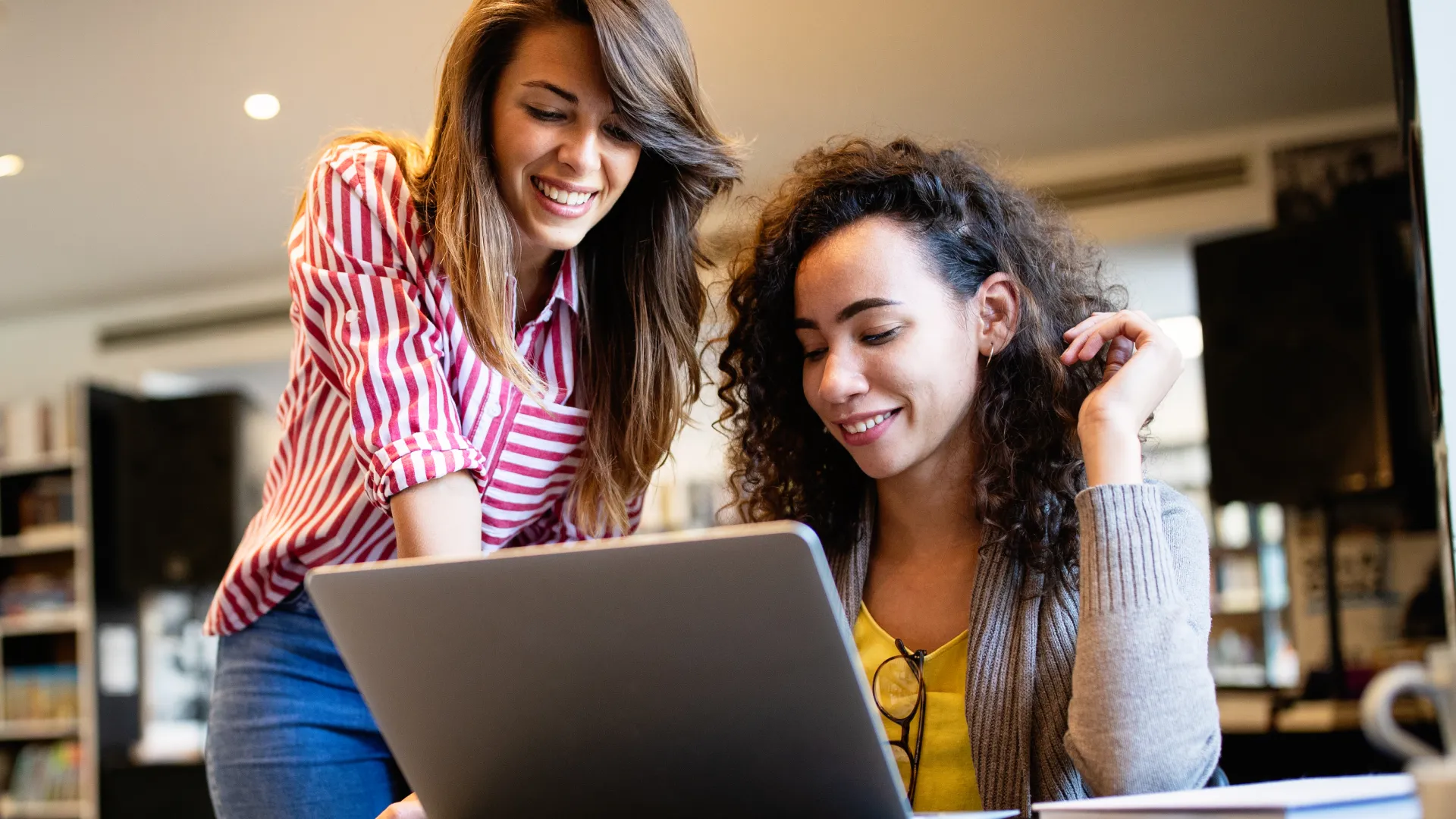 Duas estudantes universitárias colaborando em um projeto acadêmico no notebook em um ambiente de biblioteca ou sala de estudo