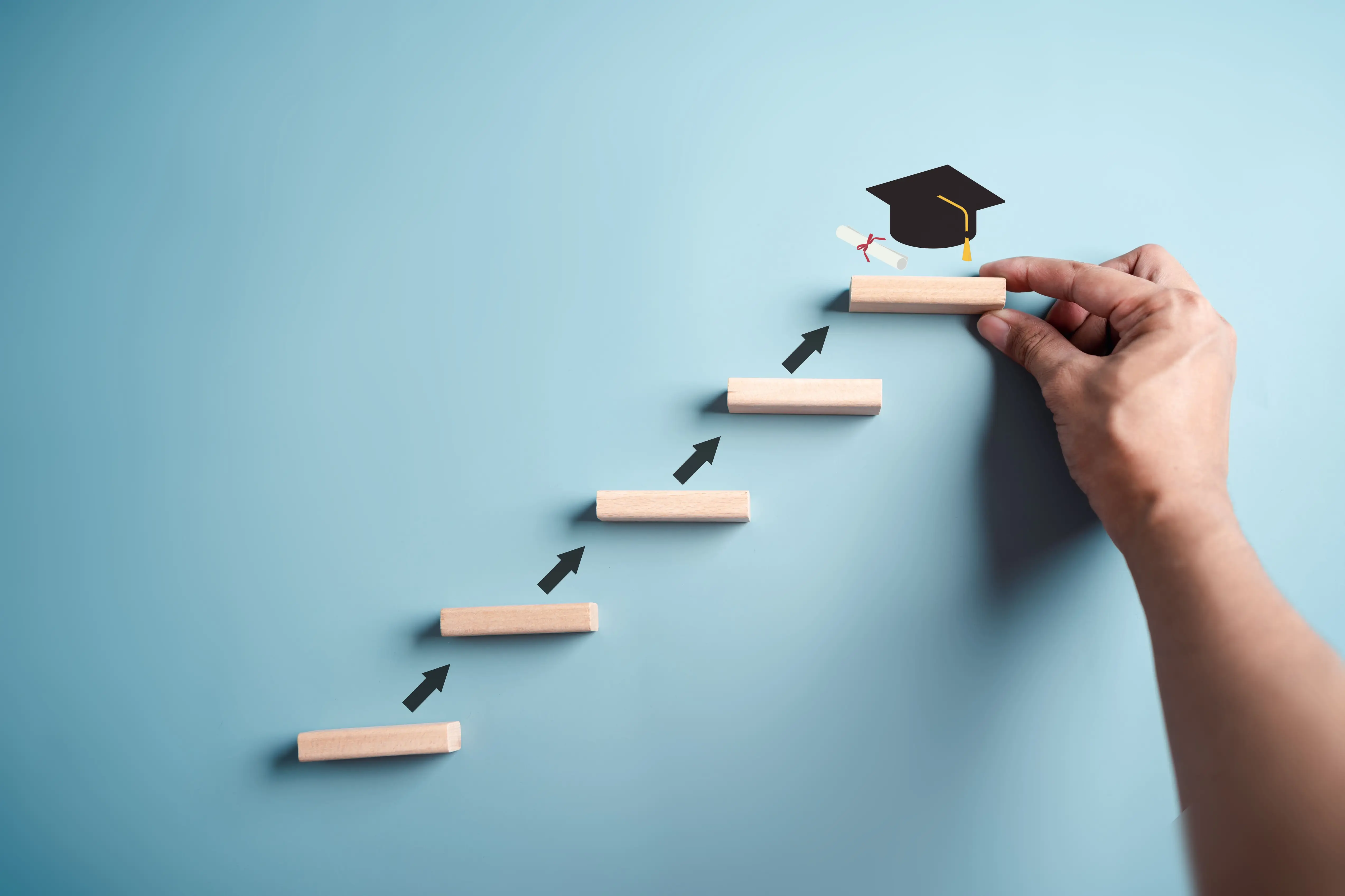 Mão montando escada de blocos de madeira com ícone de capelo de formatura no topo Conquiste seu diploma: a jornada de crescimento acadêmico e profissional através do ensino superior